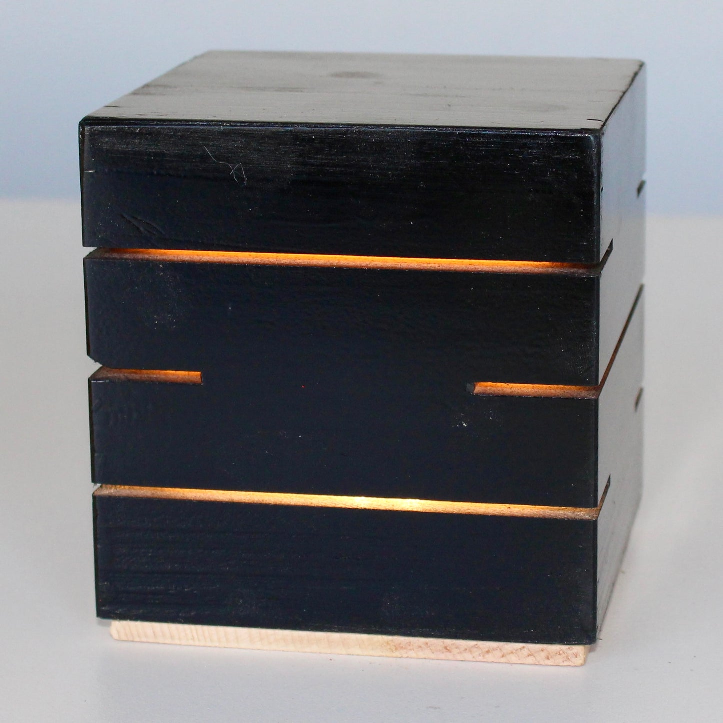 A detailed close-up, three-quarter view of the Black Cubenheim Accent size (4-inch) wood cube light from The Urbanist Collection. Handcrafted from solid wood and finished in a deep black stain, the minimalist architectural lamp glows intensely with a warm orange-gold light through precise horizontal slits. The light base shows a natural wood finish. This perspective emphasizes its clean geometric lines and brutalist-inspired aesthetic, making it a perfect sculptural centerpiece for curated modern interiors.