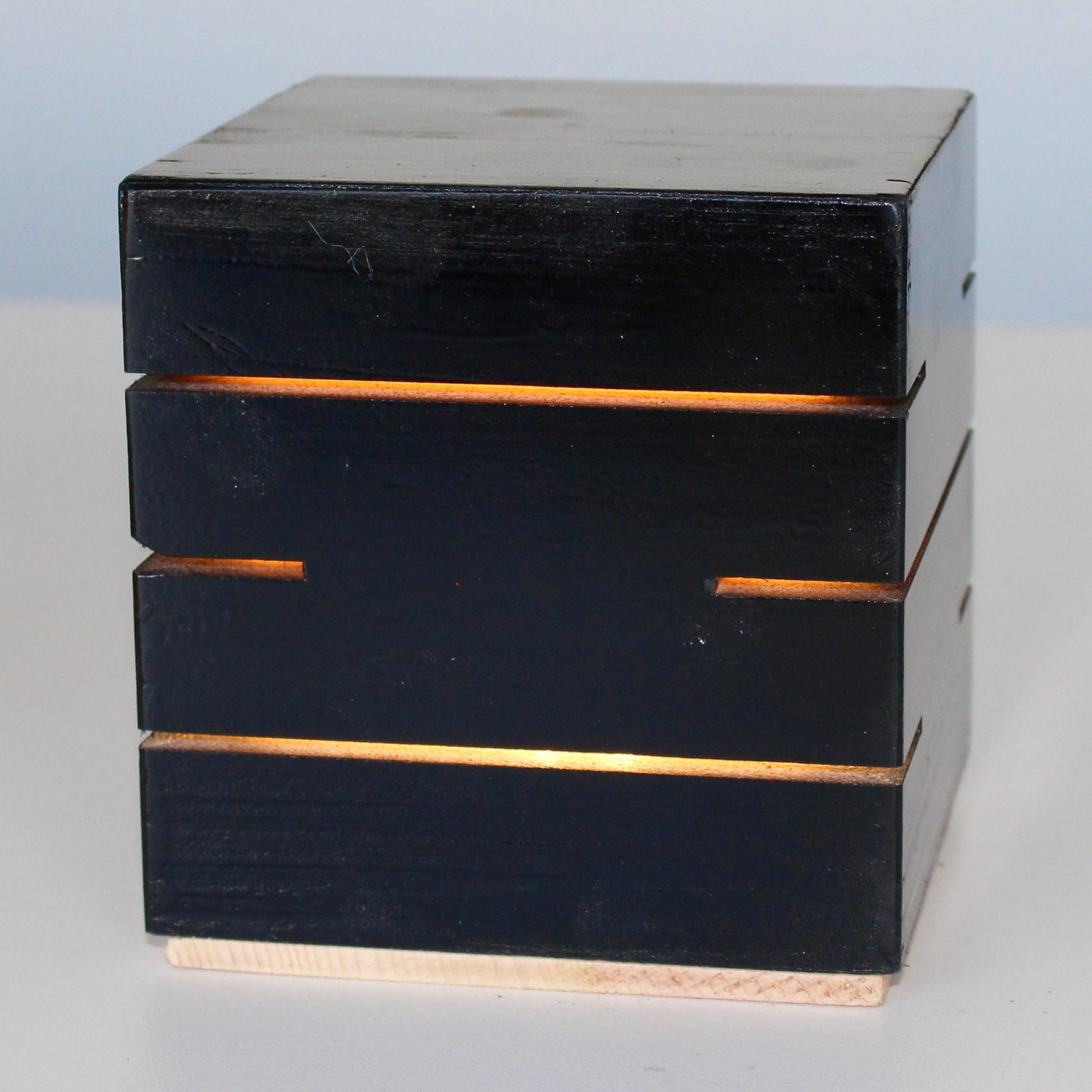 A detailed close-up, three-quarter view of the Black Cubenheim Accent size (4-inch) wood cube light from The Urbanist Collection. Handcrafted from solid wood and finished in a deep black stain, the minimalist architectural lamp glows intensely with a warm orange-gold light through precise horizontal slits. The light base shows a natural wood finish. This perspective emphasizes its clean geometric lines and brutalist-inspired aesthetic, making it a perfect sculptural centerpiece for curated modern interiors.