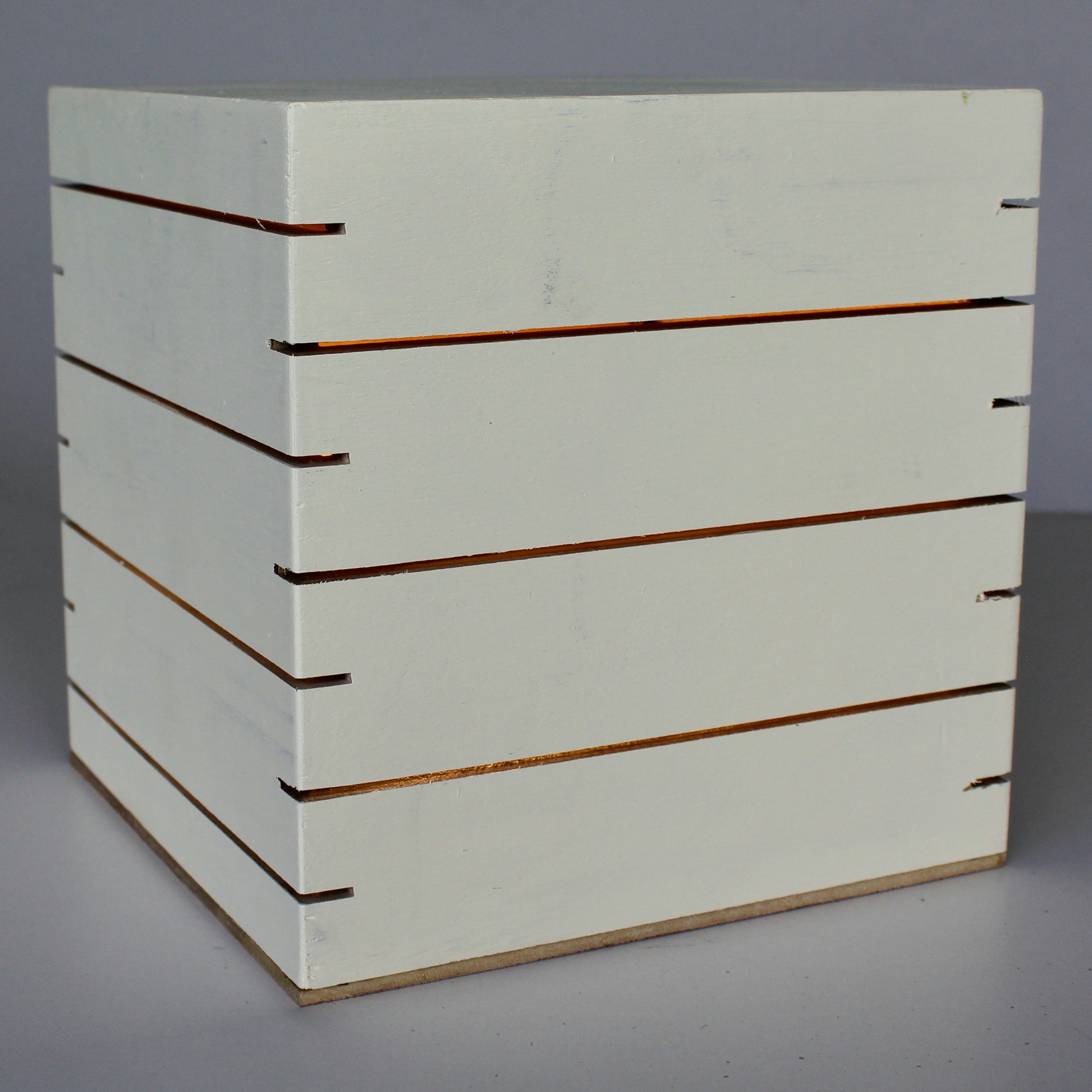 A three-quarter view of 'The Cubenheim' horizontal slit cube light from The Urbanist Series, handcrafted from solid wood with a creamy off-white Butter finish. The illuminated geometric monolith softly emits a warm orange-gold glow through multiple precisely cut horizontal vents that wrap around the block. This elegant minimalist architectural look serves as an ideal sculptural accent for mid-century modern or contemporary spaces. Natural wood base detail.