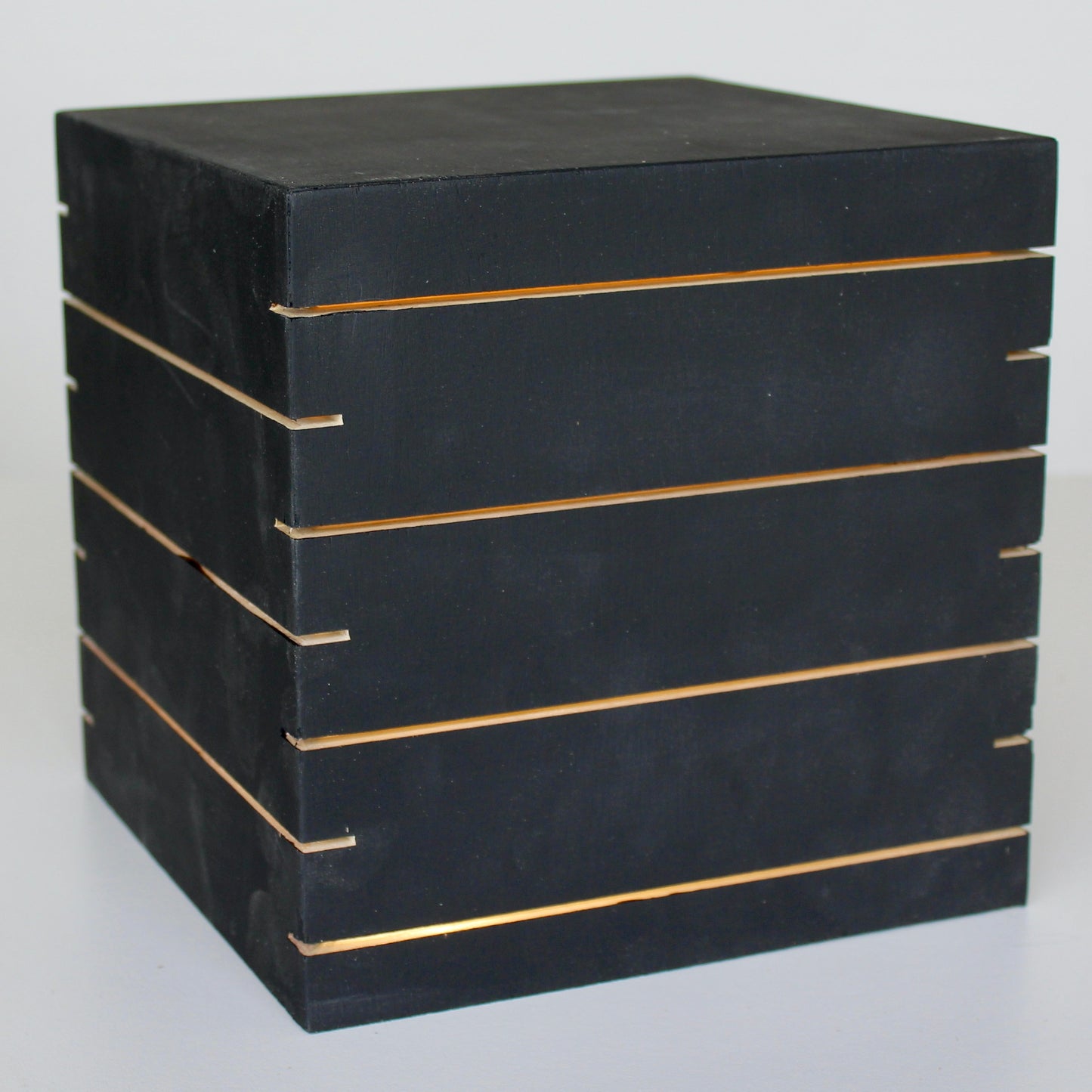 A three-quarter view of 'The Cubenheim' horizontal slit cube light from The Urbanist Series, handcrafted from solid wood with a deep black finish. The illuminated geometric monolith emits a strong, warm orange-gold glow through multiple precisely cut horizontal vents that wrap around the block. This perspective emphasizes a sleek, brutalist-inspired sculptural centerpiece. Perfect for modern, sophisticated interiors. Natural wood base detail.