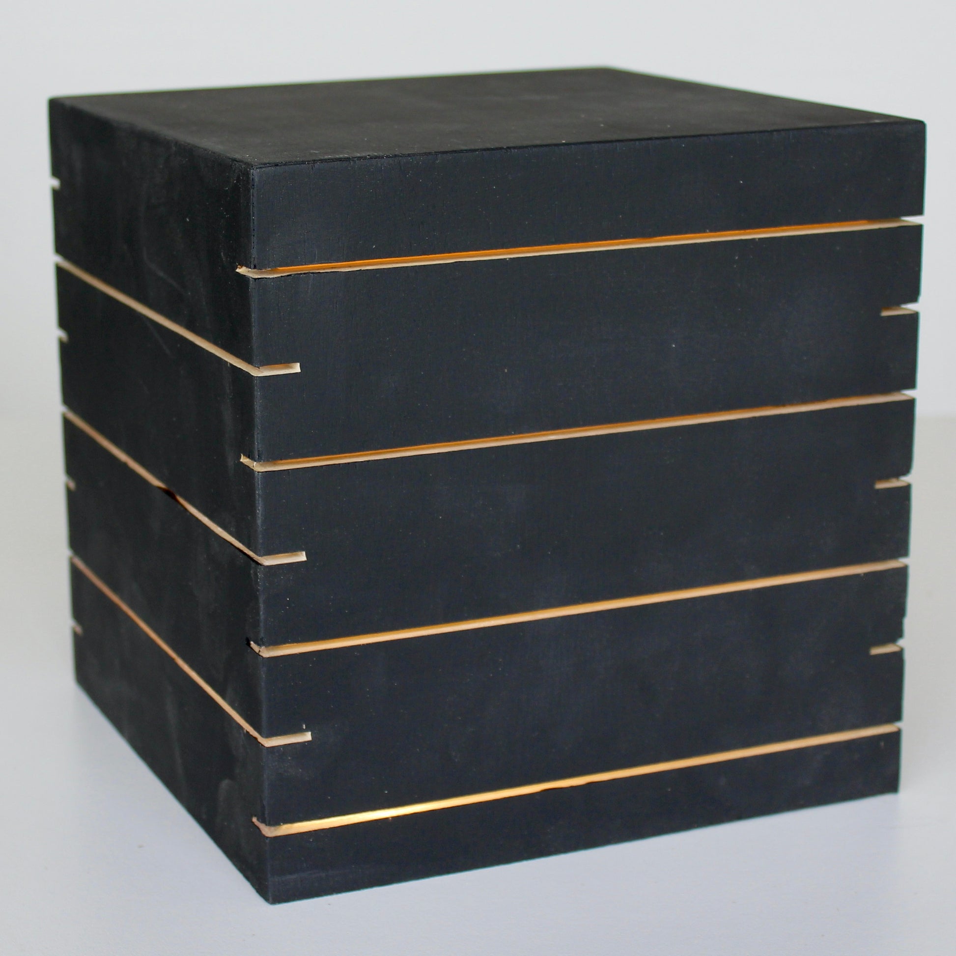 A three-quarter view of 'The Cubenheim' horizontal slit cube light from The Urbanist Series, handcrafted from solid wood with a deep black finish. The illuminated geometric monolith emits a strong, warm orange-gold glow through multiple precisely cut horizontal vents that wrap around the block. This perspective emphasizes a sleek, brutalist-inspired sculptural centerpiece. Perfect for modern, sophisticated interiors. Natural wood base detail.