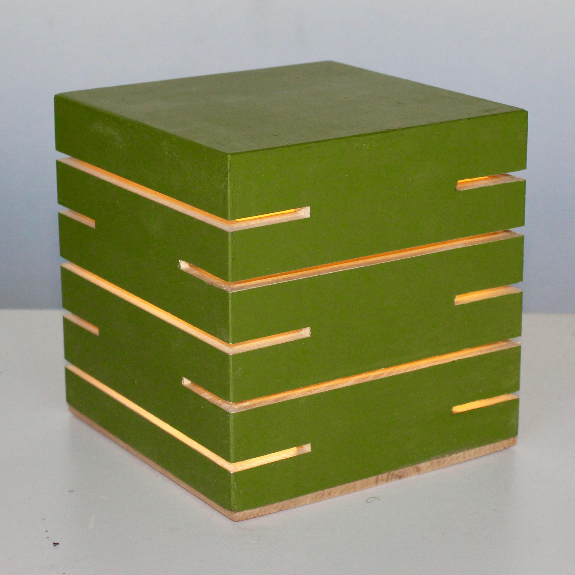 A three-quarter view of 'The Cubenheim' horizontal slit cube light in the Heirloom Avocado finish from The Urbanist Series. Handcrafted from solid wood with a sophisticated matte retro-green finish, the geometric lamp is illuminated, emitting a warm orange-gold glow from its precisely cut horizontal slots. The natural wood base provides a subtle organic contrast. This piece embodies a mid-century modern aesthetic, making it an ideal sculptural lighting accent for a home office, credenza, or bedside table.