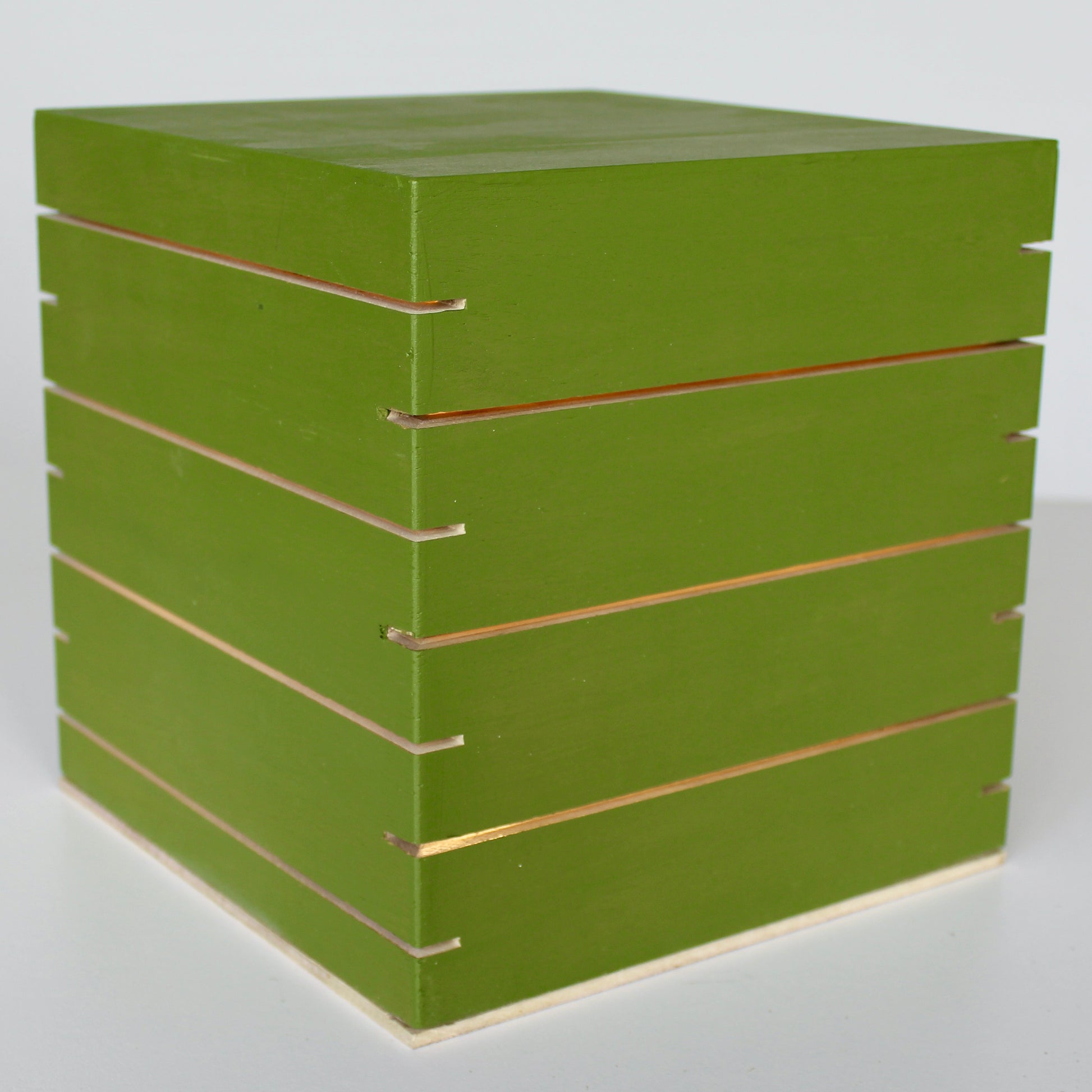 A three-quarter view of 'The Cubenheim' horizontal slit cube light from The Urbanist Series, handcrafted from solid wood with a matte retro-green Avocado finish. The illuminated geometric monolith emits a warm orange-gold glow through multiple precisely cut horizontal vents that wrap around the block. This piece embodies a distinct mid-century modern (MCM) aesthetic. A unique sculptural centerpiece for curated retro-cool living areas. Natural wood base detail.