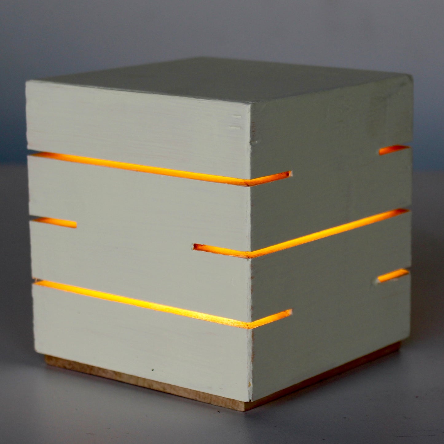 A three-quarter view photograph of 'The Cubenheim' horizontal slit wood cube light from The Urbanist Series, shown illuminated. Handcrafted from solid wood and finished in a smooth, warm, creamy Saffron Butter color stain. The minimalist geometric lamp emits a soft, warm orange-gold light through precisely cut horizontal slots, creating distinct linear light patterns. The natural wood base offers a subtle, organic contrast to the sophisticated color finish. This piece serves as a perfect sculptural lighting