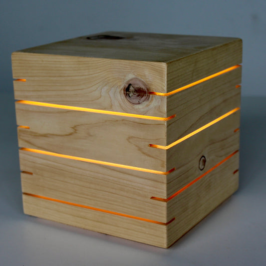 A direct 3/4 view photograph of 'The Cubenheim' horizontal slit cube light from The Urbanist Series, handcrafted from natural, light-grained cedar wood. The geometric, cuboid lamp is illuminated, emitting a warm, radiant orange-gold light from multiple precisely-cut, linear horizontal slits that wrap around its form. This perspective highlights the wood's prominent grain and a distinctive natural knot, showcasing a brutalist, architectural minimalist aesthetic. The lamp is resting on a clean studio surface.