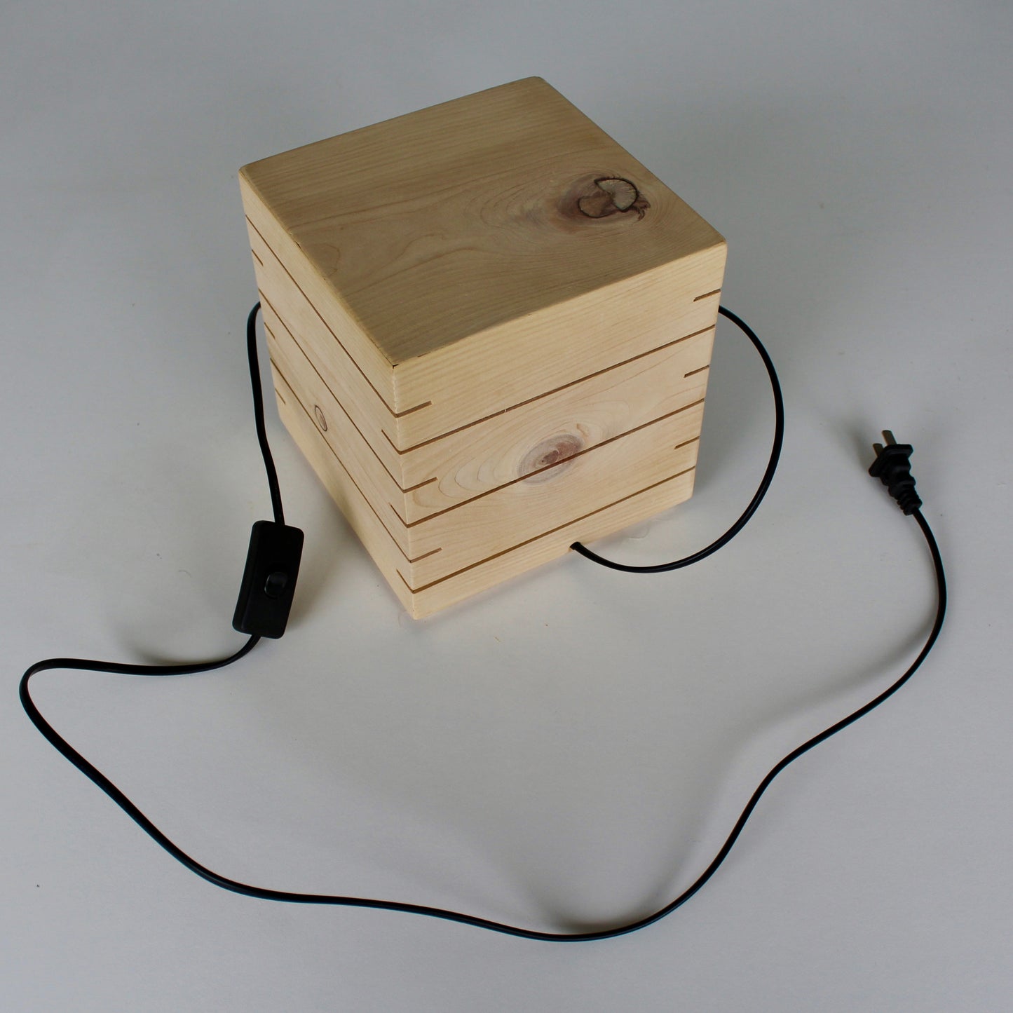 A clean, high-angle overview photograph of 'The Cubenheim' horizontal slit cube light from The Urbanist Series, shown in its unlit state. The geometric lamp is handcrafted from light-grained natural cedar wood, featuring multiple precise unlit horizontal slots on the visible top and front-side faces. A long black electrical cord with a visible inline thumb switch and standard power plug extends from the rear-side base and rests on the clean white surface, detailing the complete product set. The perspective 