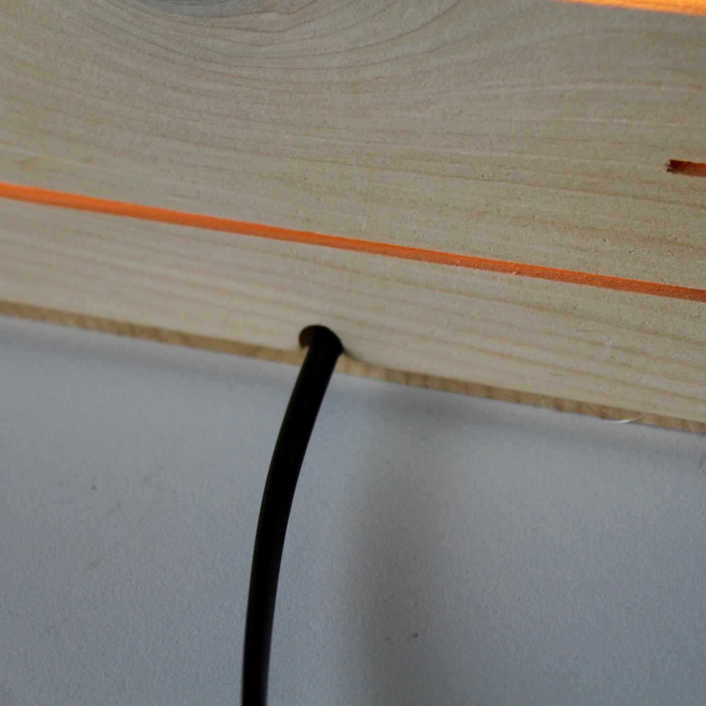 A close-up, rear-view detail of 'The Cubenheim' horizontal slit cube light from The Urbanist Series. The photograph highlights the clean craftsmanship of the handcrafted natural cedar body, featuring a black power cord exiting from a precisely carved notch at the base. Above the cord, a single horizontal slit glows with a warm orange-gold light, showcasing the wood's fine grain and the seamless integration of electrical components in this minimalist architectural luminaire.