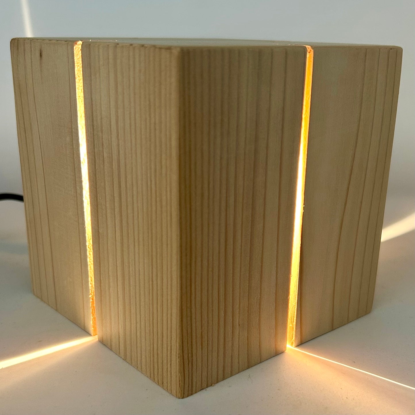 A detailed close-up, three-quarter view of 'The Salk' vertical slit cube light, showcasing the rich, intricate texture of the light-colored natural cedar wood grain. The handcrafted wood cube is illuminated from within, with a warm light glowing brightly from the precise vertical slits and casting a crisp line of projected light across the white surface below.