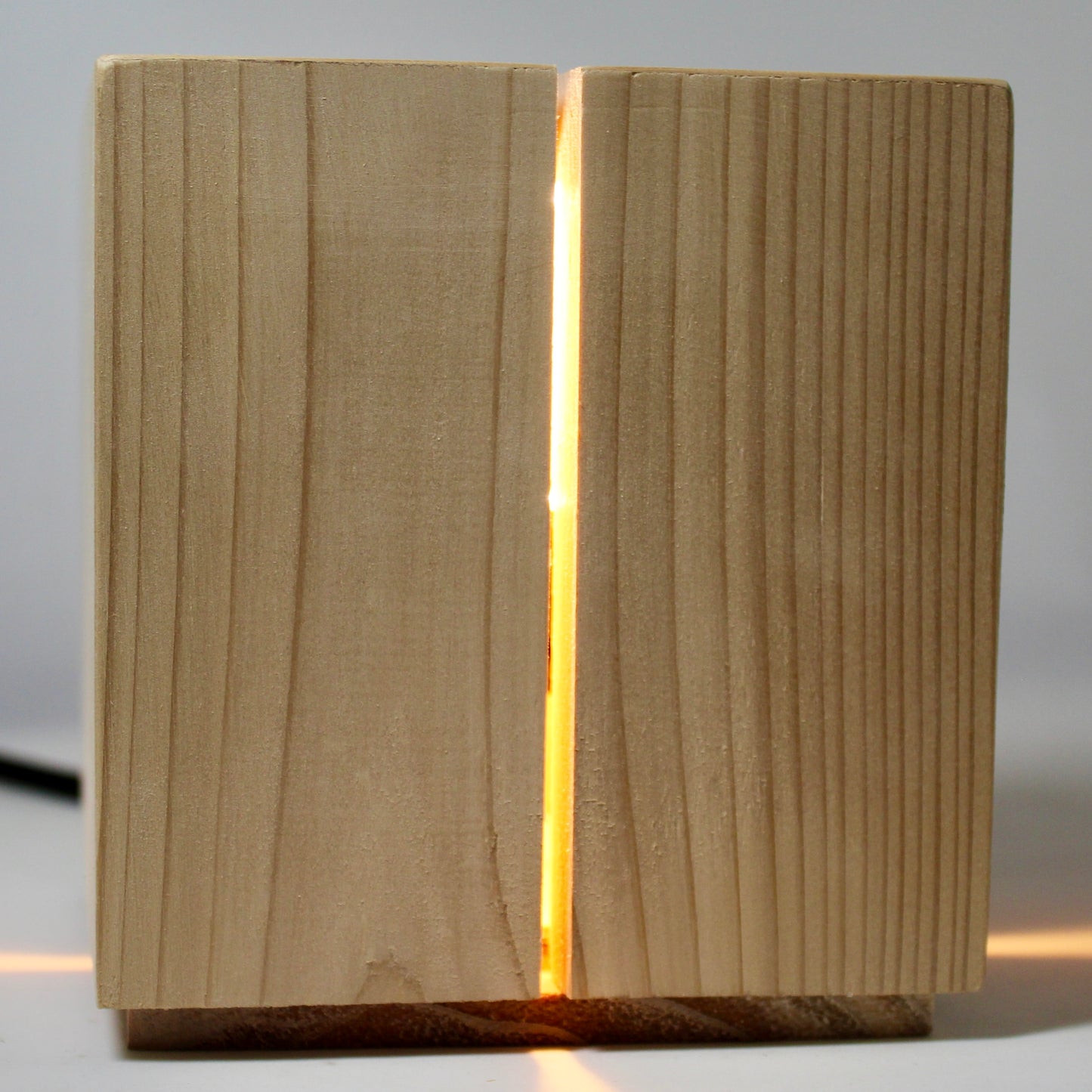 A powerful monolithic front view of 'The Salk' vertical slit cube light from The Urbanist Series, handcrafted from natural cedar wood. The geometric lamp stands on a light grey surface, with a warm, radiant orange-gold light beaming out intensely from the precise vertical slits and casting distinct architectural shadows. This perspective highlights the vertical rhythm and clean lines of the minimalist design, making the solid cedar block appear to glow with internal life.