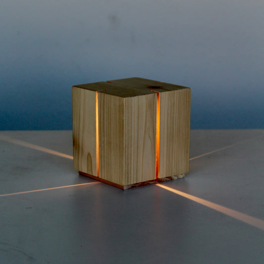 A three-quarter view photograph of 'The Salk' vertical slit cube light, handcrafted from light-grained natural cedar wood. The lamp is sitting on a gray studio surface and is illuminated, casting a warm orange glow. Two precise vertical slits are visible on the front and side faces, and the light projects a dramatic, distinct cross-shaped pattern of light beams onto the surface beneath it.