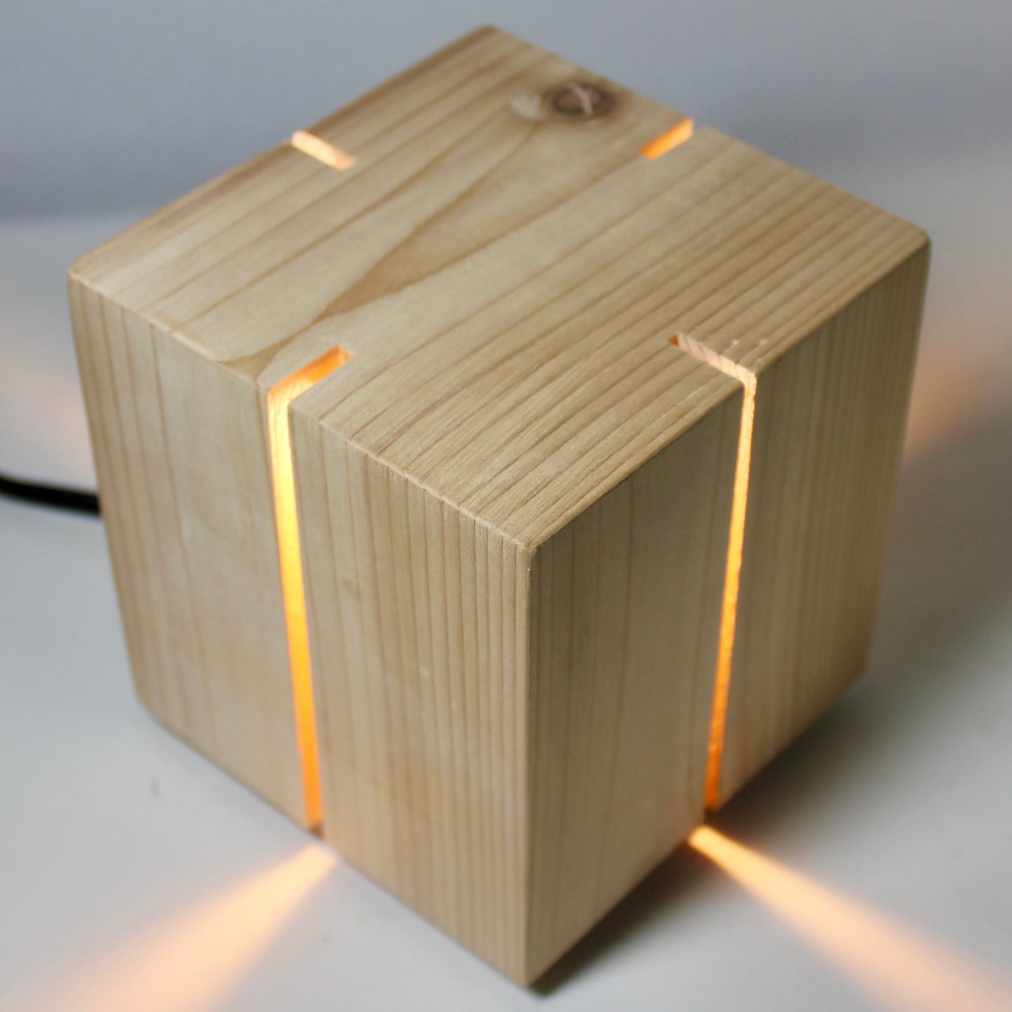 A high-angle, birds-eye view of 'The Salk' vertical slit cube light from The Urbanist Series. Handcrafted from solid natural cedar, the top of the cube shows unique wood grain and a natural knot detail. Warm light glows through the four vertical corner slits, projecting four distinct, symmetrical beams of light outward across the white studio surface. The piece exemplifies a minimalist, architectural aesthetic with its clean geometric lines.