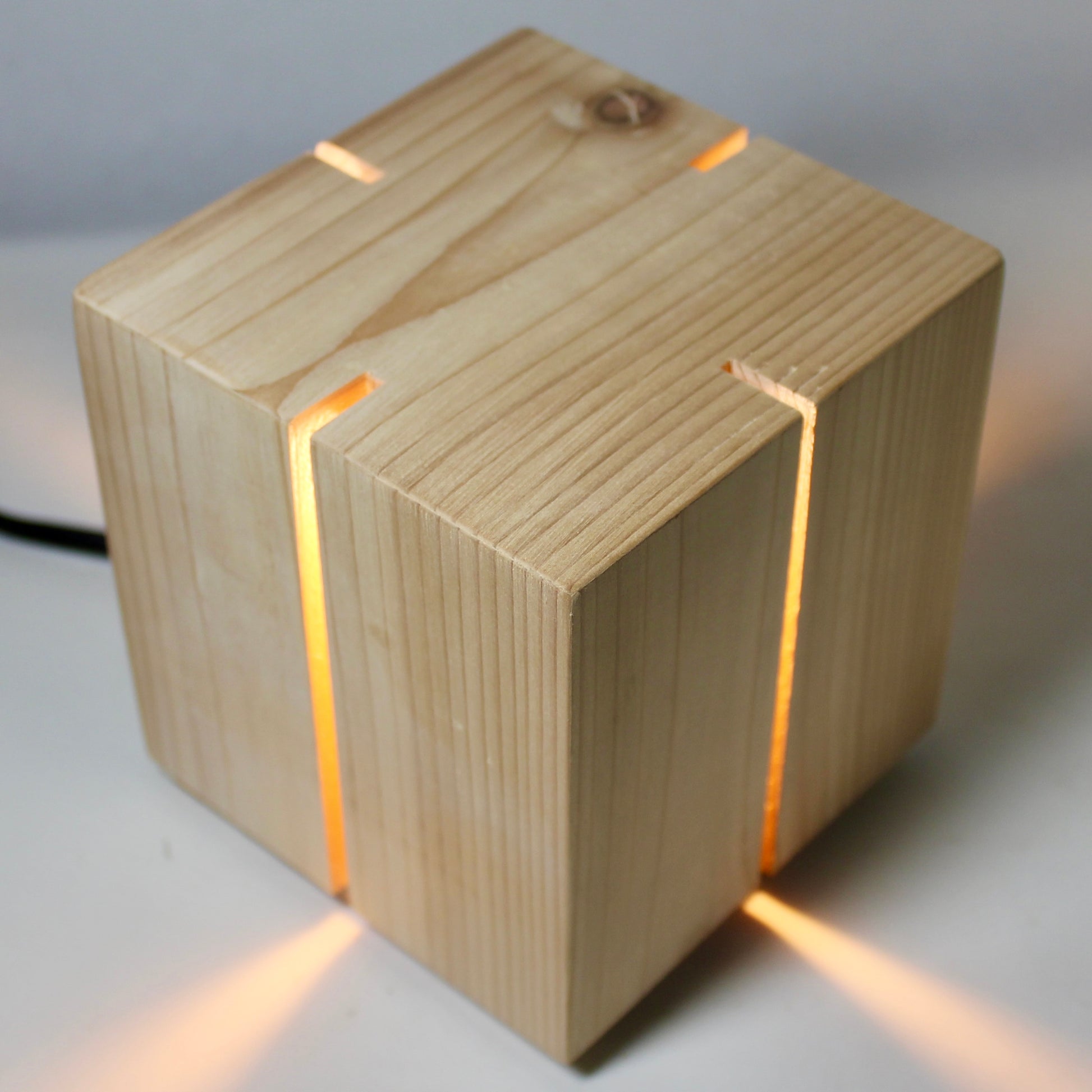 A high-angle, birds-eye view of 'The Salk' vertical slit cube light from The Urbanist Series. Handcrafted from solid natural cedar, the top of the cube shows unique wood grain and a natural knot detail. Warm light glows through the four vertical corner slits, projecting four distinct, symmetrical beams of light outward across the white studio surface. The piece exemplifies a minimalist, architectural aesthetic with its clean geometric lines.