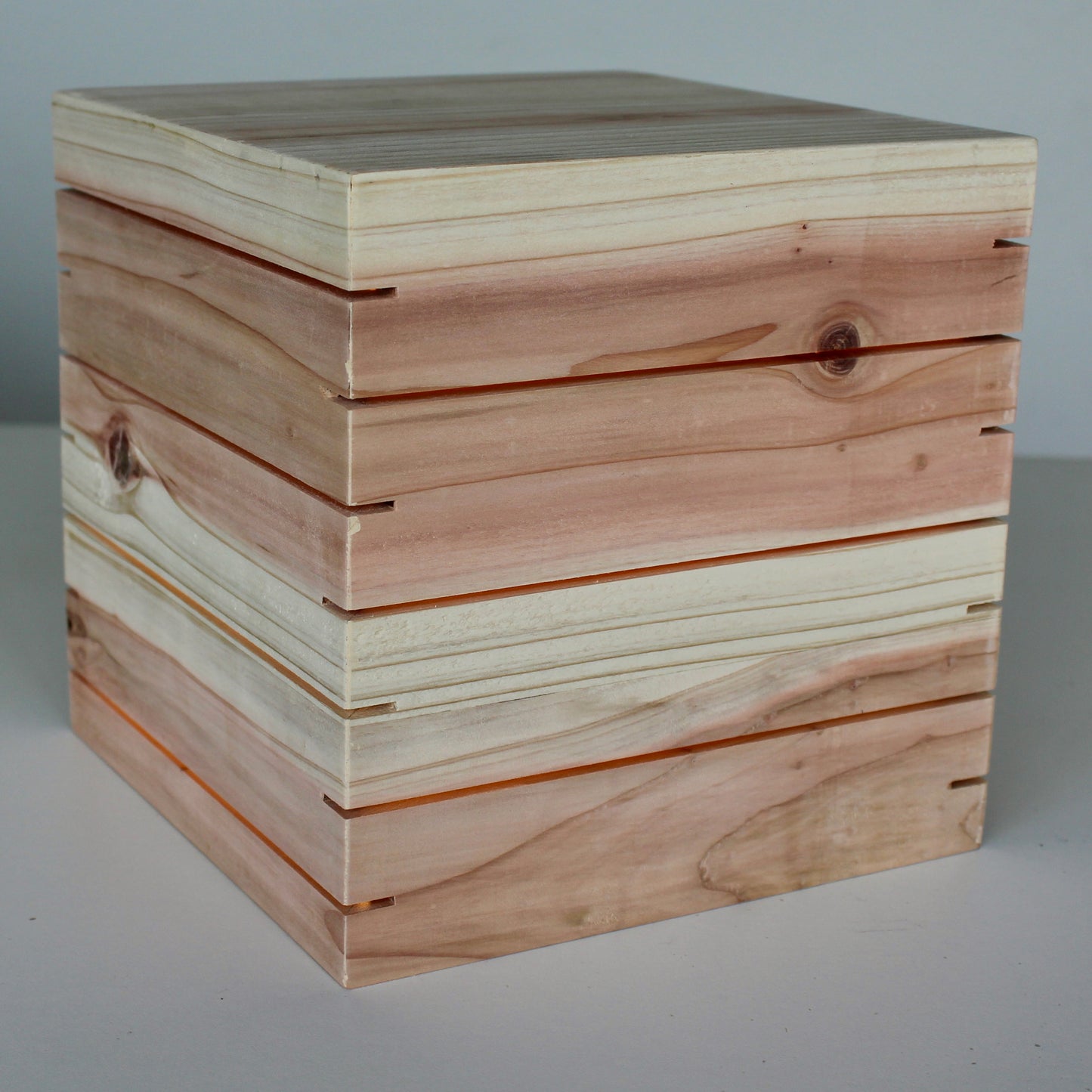An unlit three-quarter view of 'The Cubenheim' horizontal slit cube sculpture from The Urbanist Series. Handcrafted from premium natural cedar, this non-illuminated perspective captures the spectacular raw grain and natural contrast between the reddish heartwood and lighter sapwood. Several prominent natural knots are visible. The precisely cut horizontal vents wrap around the geometric block, highlighting a monolithic and brutalist-inspired wood sculpture suitable for office or shelf display.